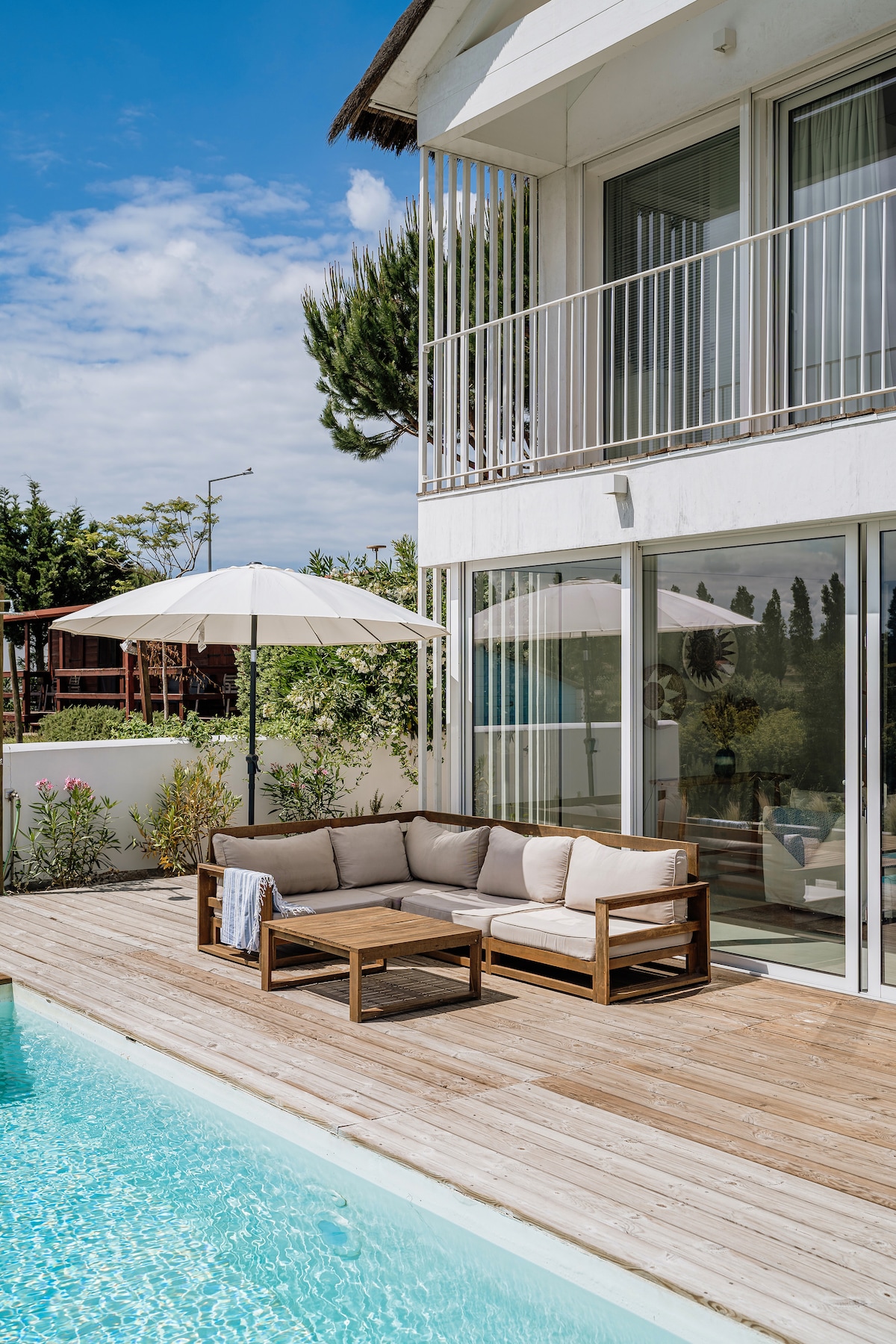 An outdoor deck features a wooden seating area with light cushions, complemented by a large umbrella. A sparkling salt-water pool lies nearby, and glass sliding doors open to the interior, allowing natural light to illuminate the space.