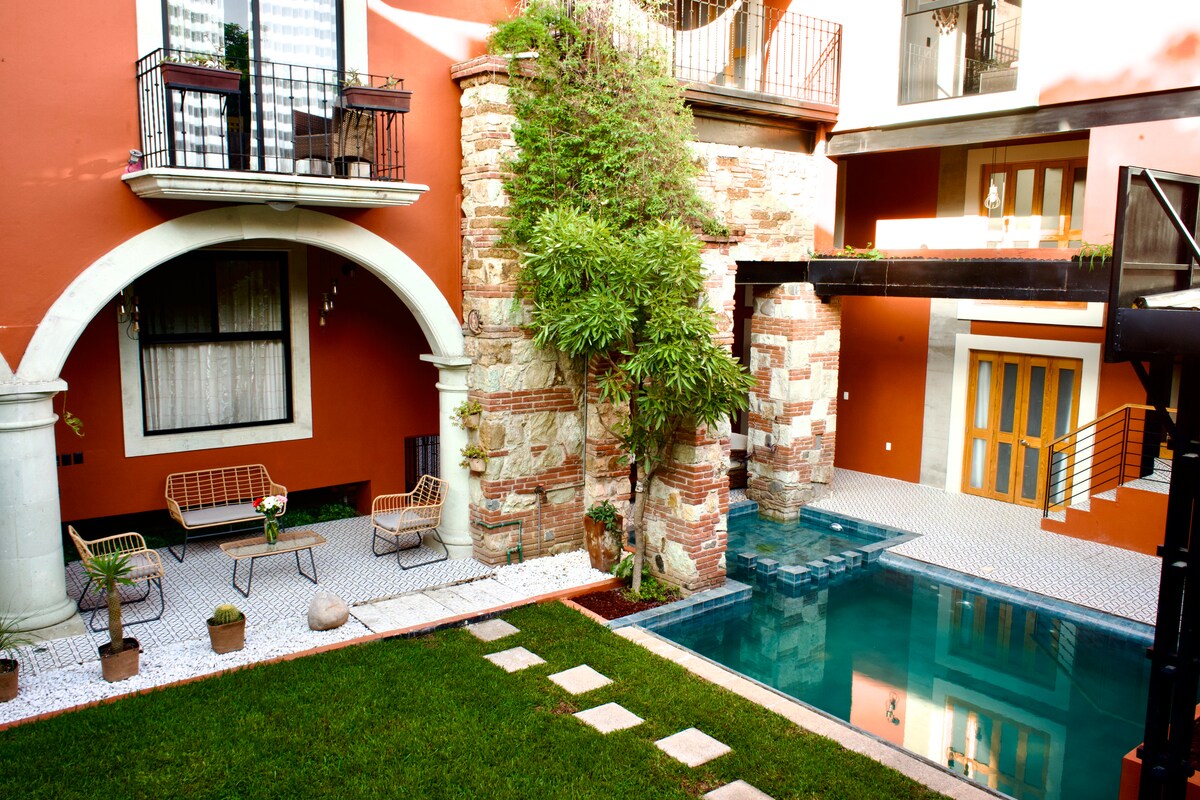 An outdoor area showcases a well-maintained garden with green grass, stone pathways, and potted plants. A pool reflects the surrounding architecture, with a cozy seating arrangement featuring a couple of metal benches. The orange walls of the building add a warm backdrop to the space.