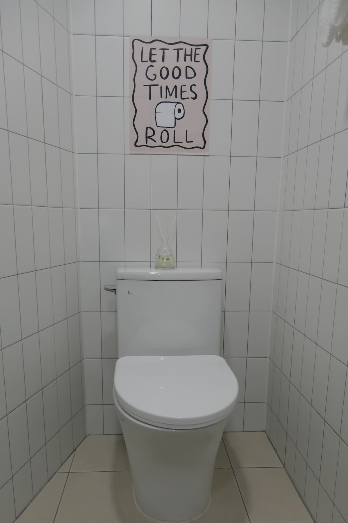 A clean and minimalistic bathroom is illustrated, featuring white tiled walls and a modern toilet. On the wall, a light-hearted sign reads 'LET THE GOOD TIMES ROLL.' A decorative diffuser sits atop the toilet, adding a subtle touch to the space.