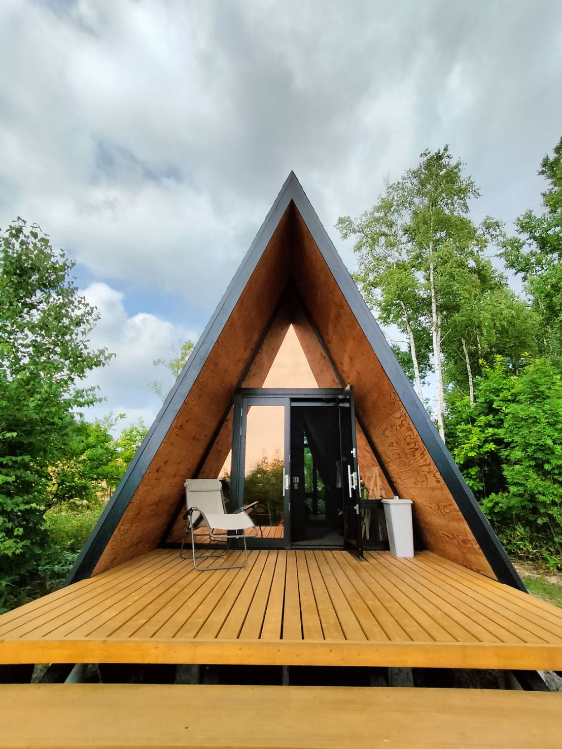 The A-Frame structure features a striking angular design and a wooden deck leading to double glass doors. Surrounded by lush greenery, a single chair is positioned on the deck, providing a space to relax and enjoy the natural setting.