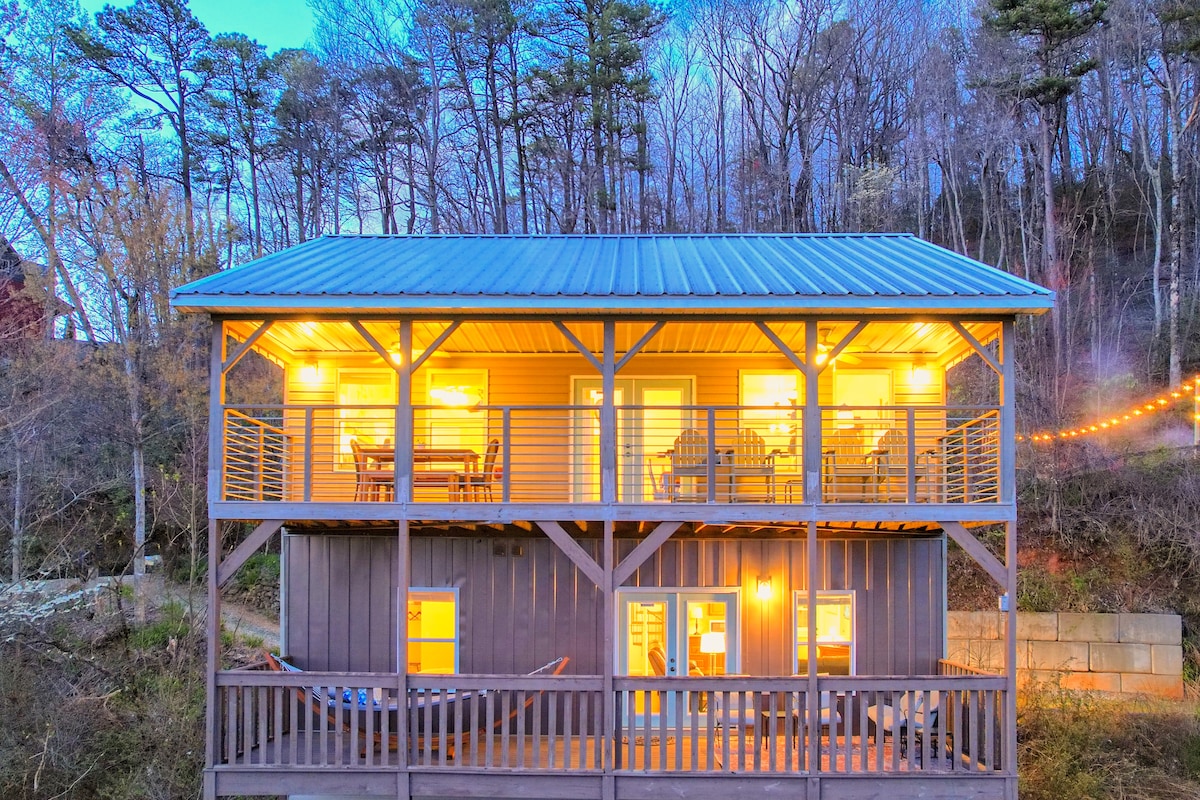 The cabin is shown from an exterior angle, highlighting two levels illuminated by warm lights. Spacious decks with seating invite relaxation, while trees line the background, contributing to the serene setting. The structured design showcases large windows, blending indoor comfort with the natural environment.