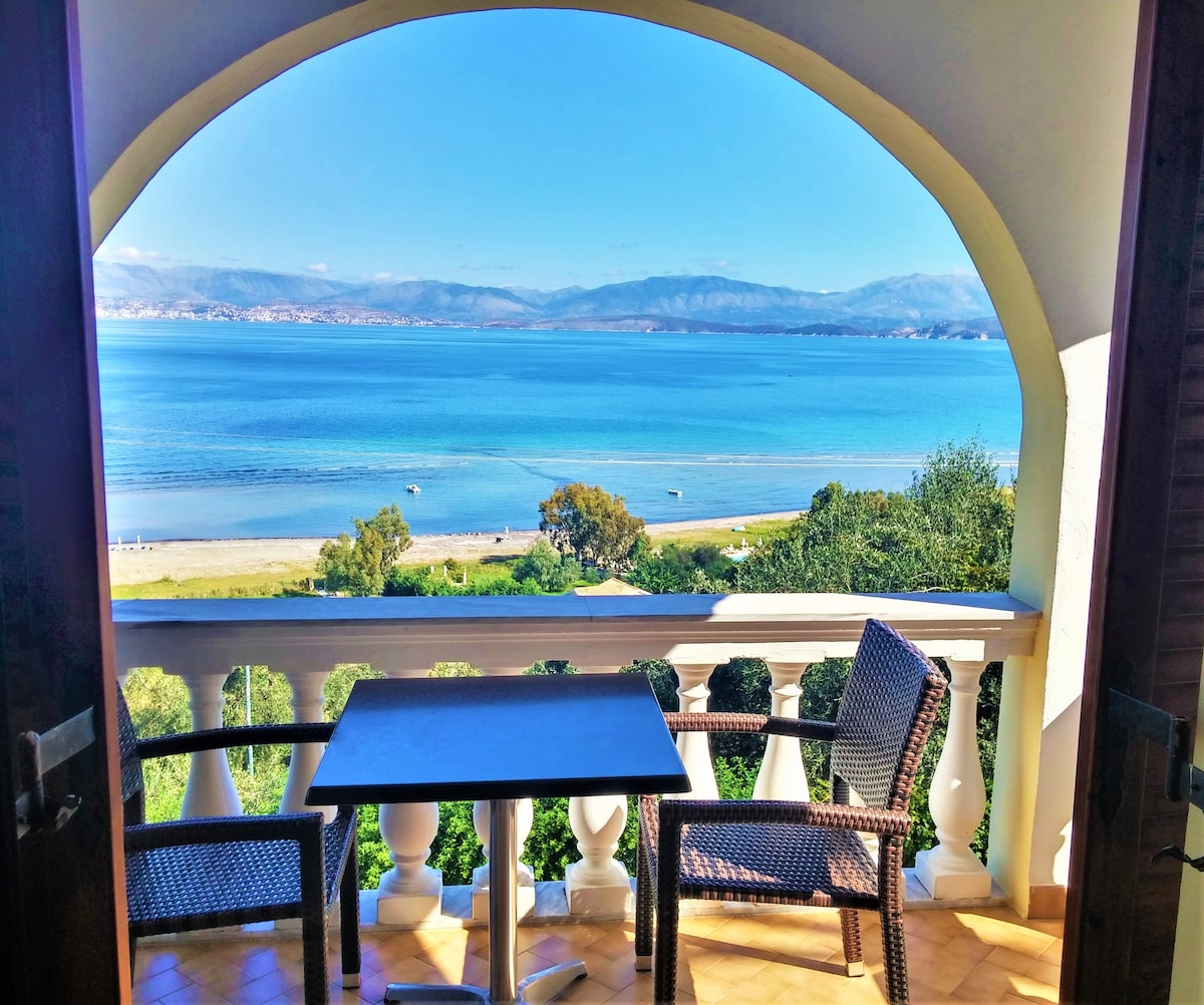 A balcony view features a table and two wicker chairs, framed by an archway. The expansive sea stretches beyond, with calm blue waters reflecting the sky. In the distance, gentle hills are visible, while the shoreline is lined with greenery and sandy beaches.