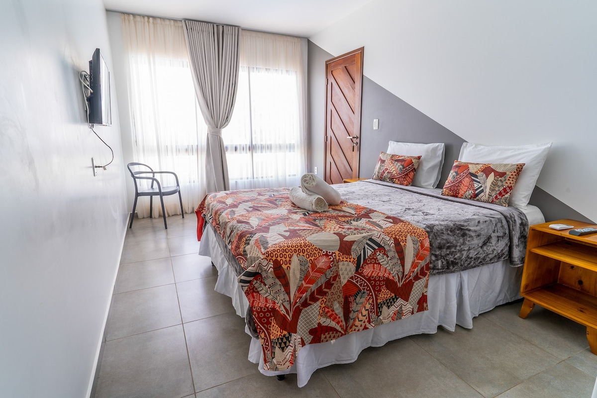 A well-appointed room features a spacious bed adorned with a colorful quilt and decorative pillows. Natural light filters through sheer curtains, illuminating the area. A small chair and a wooden bedside table complement the space, while a television is mounted on the wall.