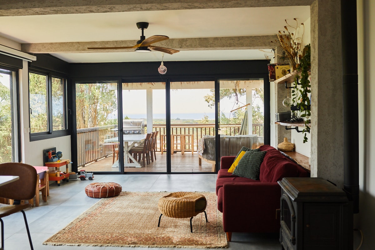 A modern living space features large glass doors that open to a balcony with views. A comfortable sofa and decorative pillows are positioned near a warm wood stove. Natural light illuminates the room, highlighting a woven rug and hints of greenery from indoor plants.