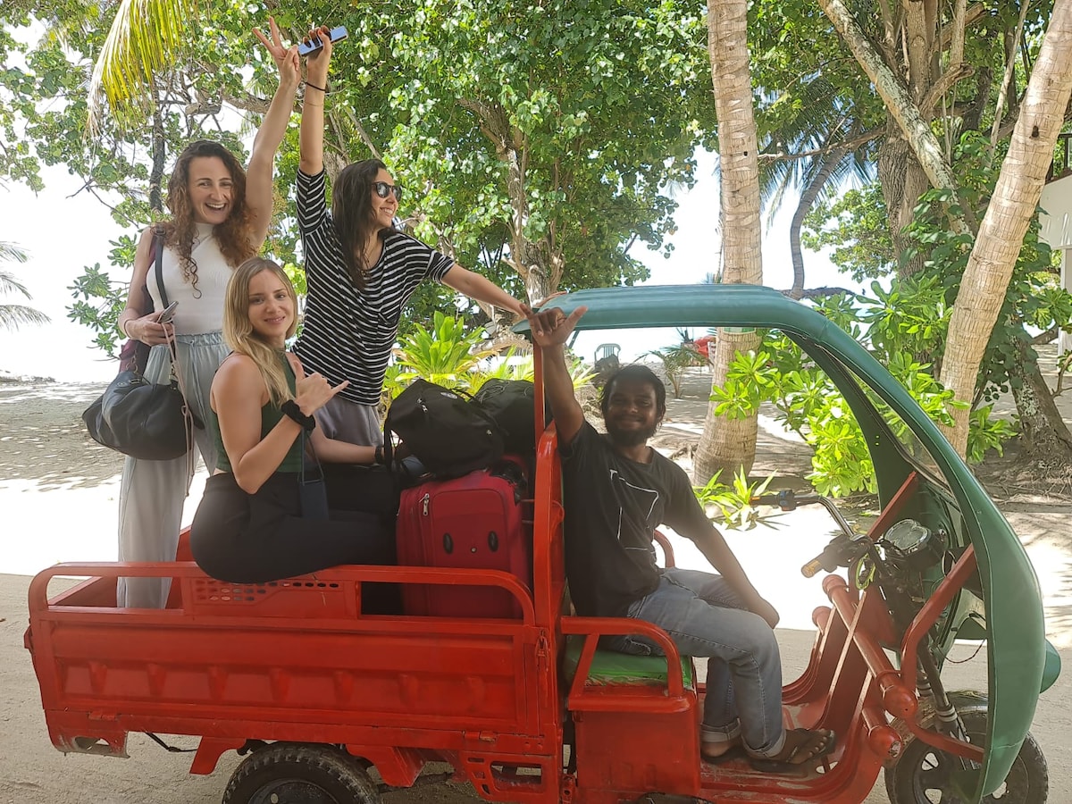 A vibrant red tuk-tuk awaits along the sandy path, carrying four individuals celebrating their arrival. Luggage is stacked at the back, while palm trees offer shade and a view of the beach in the background, enhancing the sense of adventure and relaxation.