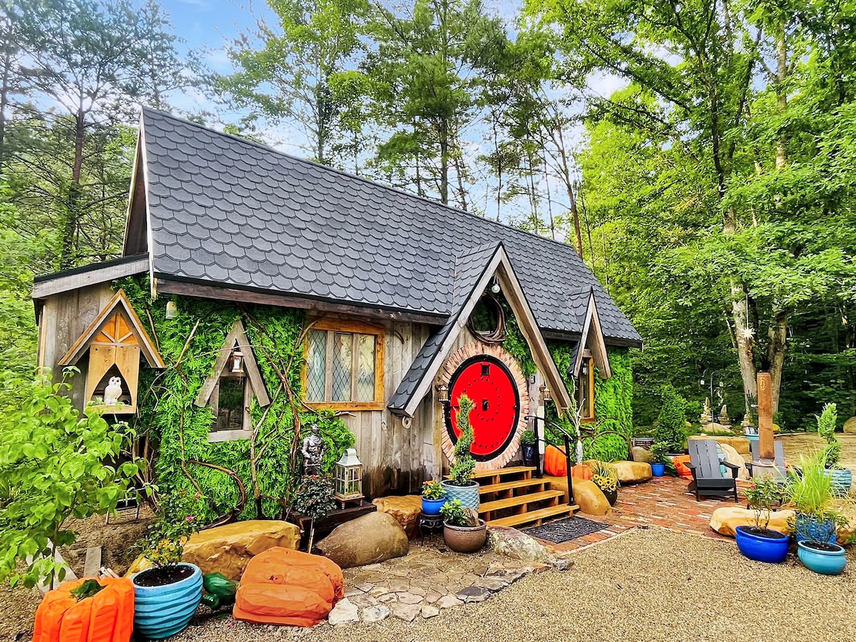 The exterior of a charming tiny house is surrounded by lush greenery and vibrant potted plants. Colorful seating options, including bean bags and smooth stones, invite relaxation. The distinctive design features a steep roof, wooden trim, and large circular doorway, contributing to the whimsical aesthetic.