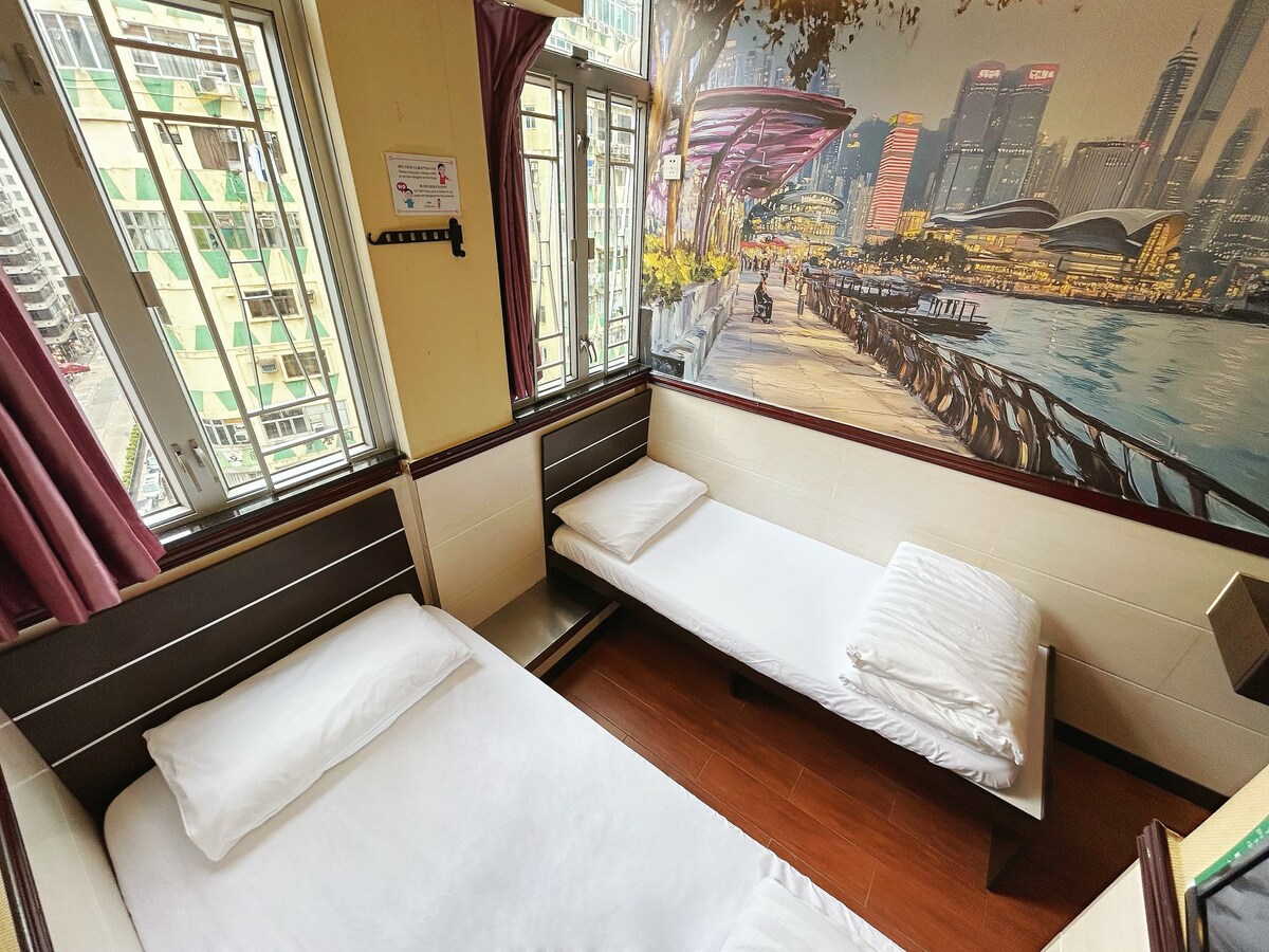 A sleeping area features two beds, one with a white linen cover and the other prepared with stacked white pillows. A large mural depicting a cityscape decorates the wall, while windows allow natural light to fill the space, framed by maroon curtains.