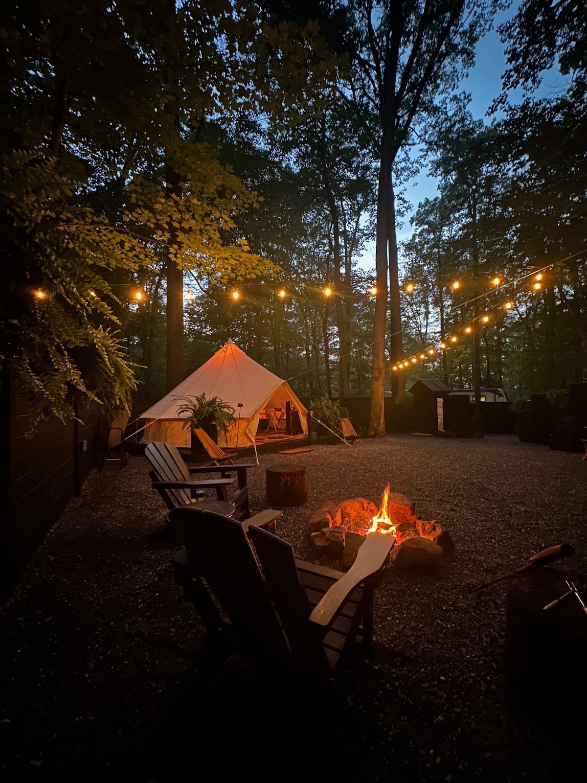 Soft lighting from string lights hangs overhead, illuminating a glamping tent nestled among trees. A fire pit with a crackling fire sits in the foreground, surrounded by rustic wooden seating. The setting offers a serene ambiance as twilight descends over the woodland area.
