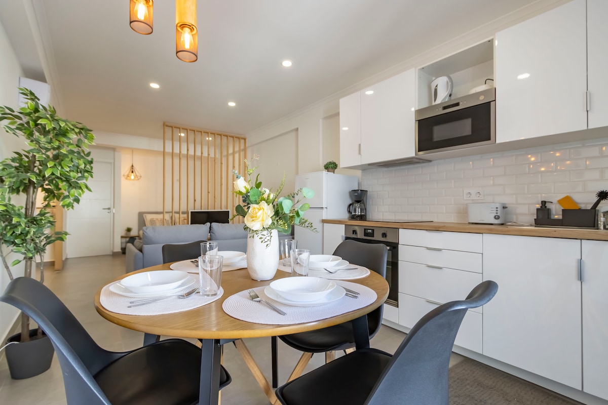A modern dining area is highlighted by a round wooden table set for four. Stylish black chairs surround the table, complemented by a vase of fresh flowers. The kitchen area, featuring sleek cabinetry and essential appliances, is visible in the background.