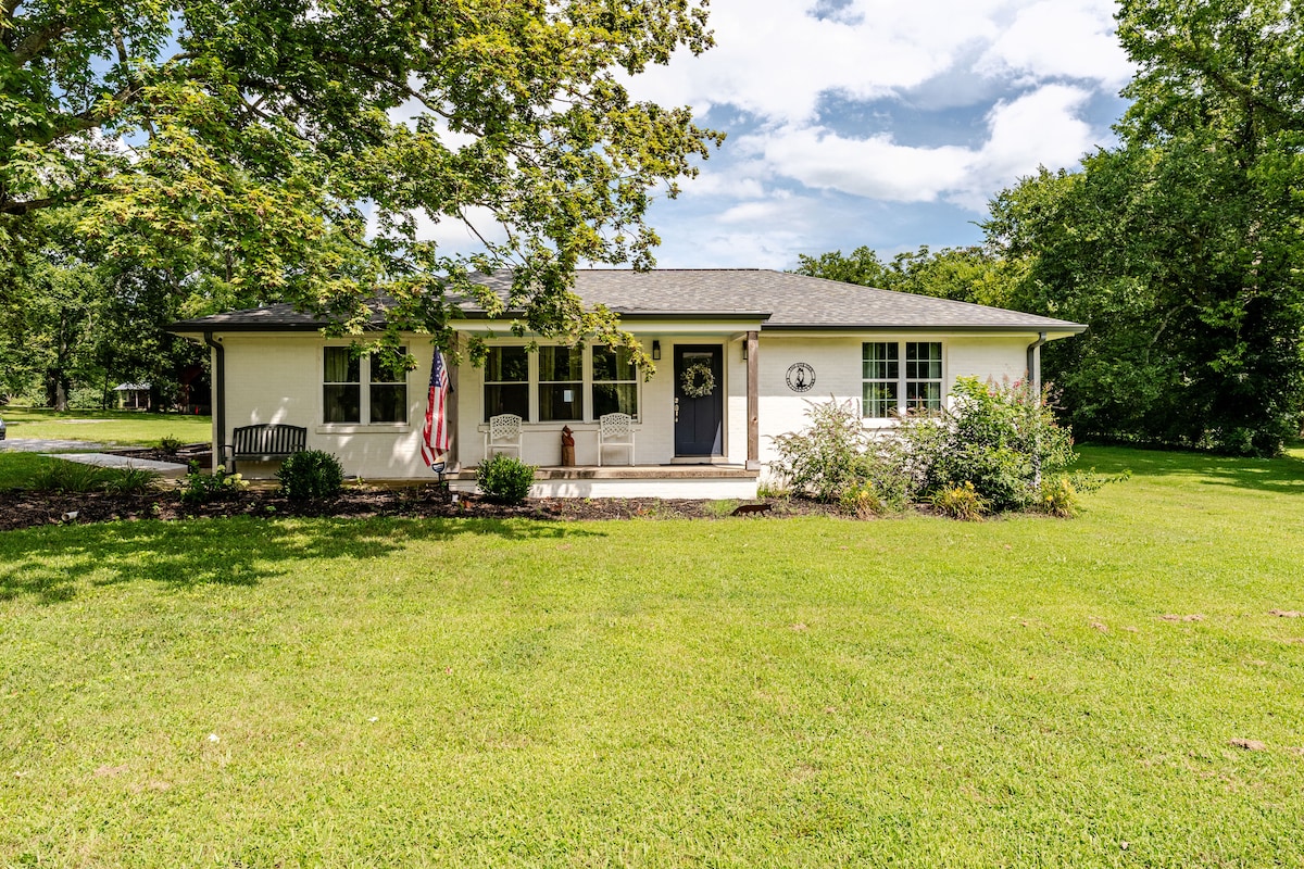 A charming farmhouse sits surrounded by lush green grass and mature trees. The front porch is adorned with a welcoming bench and a decorative wreath on the door. An American flag hangs proudly, reflecting a classic rural aesthetic.