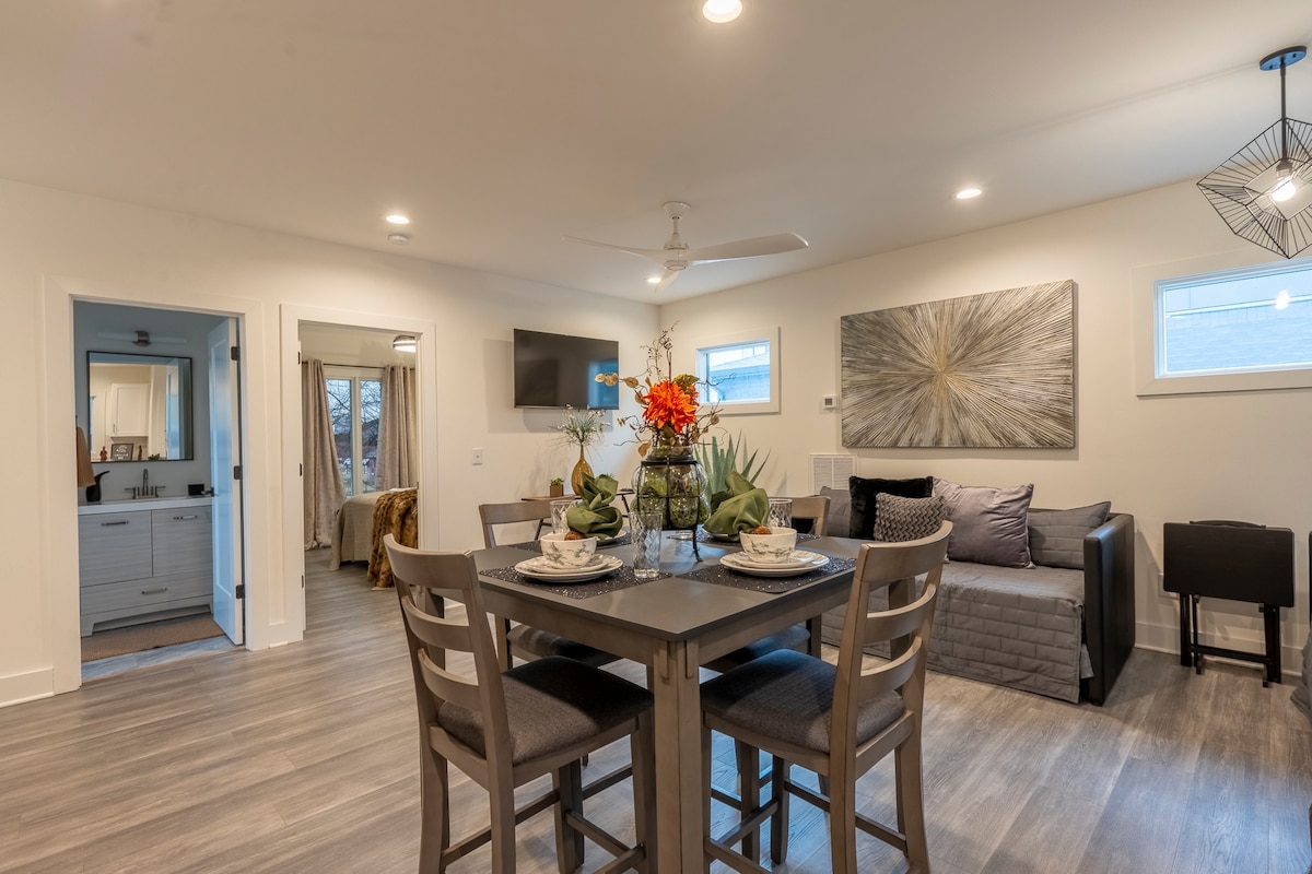 A modern dining area is highlighted by a square table surrounded by four chairs, elegantly set with dishware. A cozy seating area is visible in the background, featuring a sofa and television. Bright artwork adorns the wall, enhancing the inviting atmosphere.