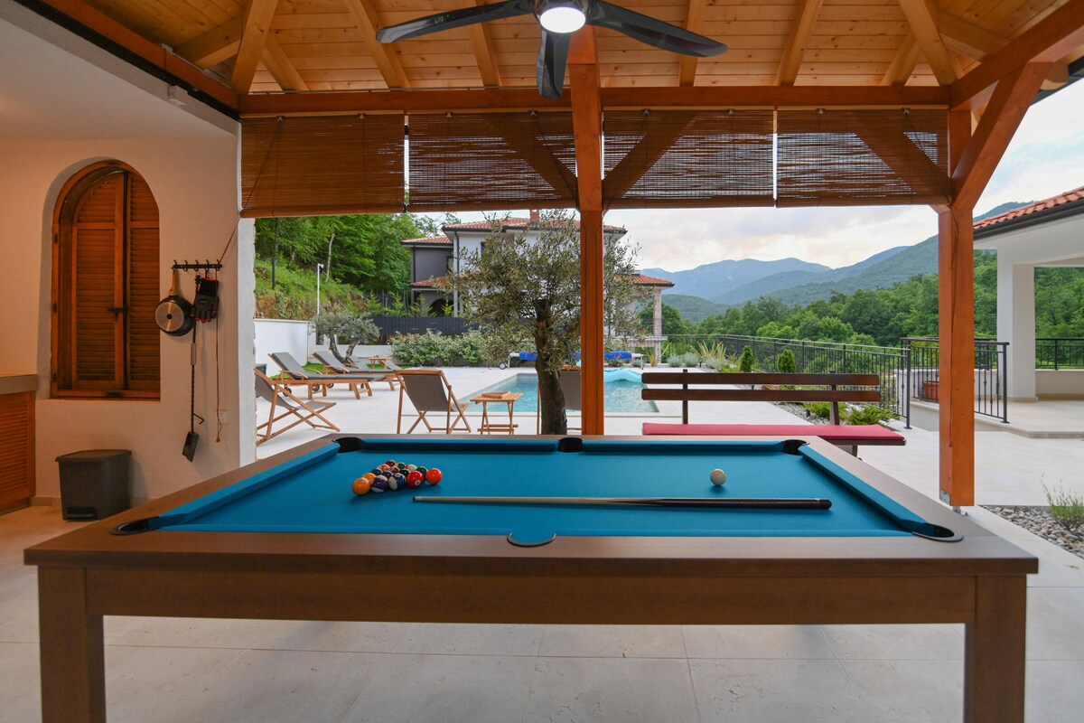 An outdoor leisure area features a billiards table under a wooden pergola. In the background, a heated pool is visible, surrounded by lounge chairs and lush greenery. Scenic mountain views are framed by large windows and a wooden structure.