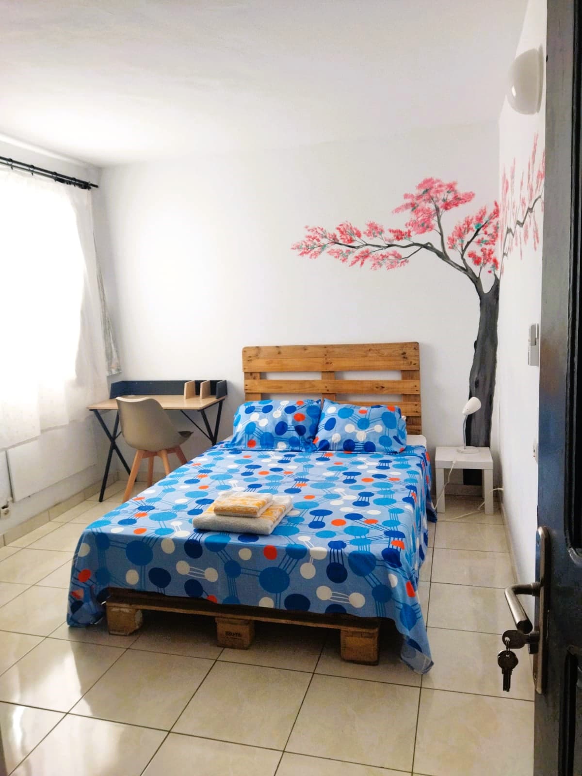 A bright and spacious room features a bed adorned with a colorful polka dot bedspread and decorative pillows. A small workspace with a chair is positioned beside the bed, and a painted cherry blossom tree decorates one wall. Natural light filters through a sheer curtain.
