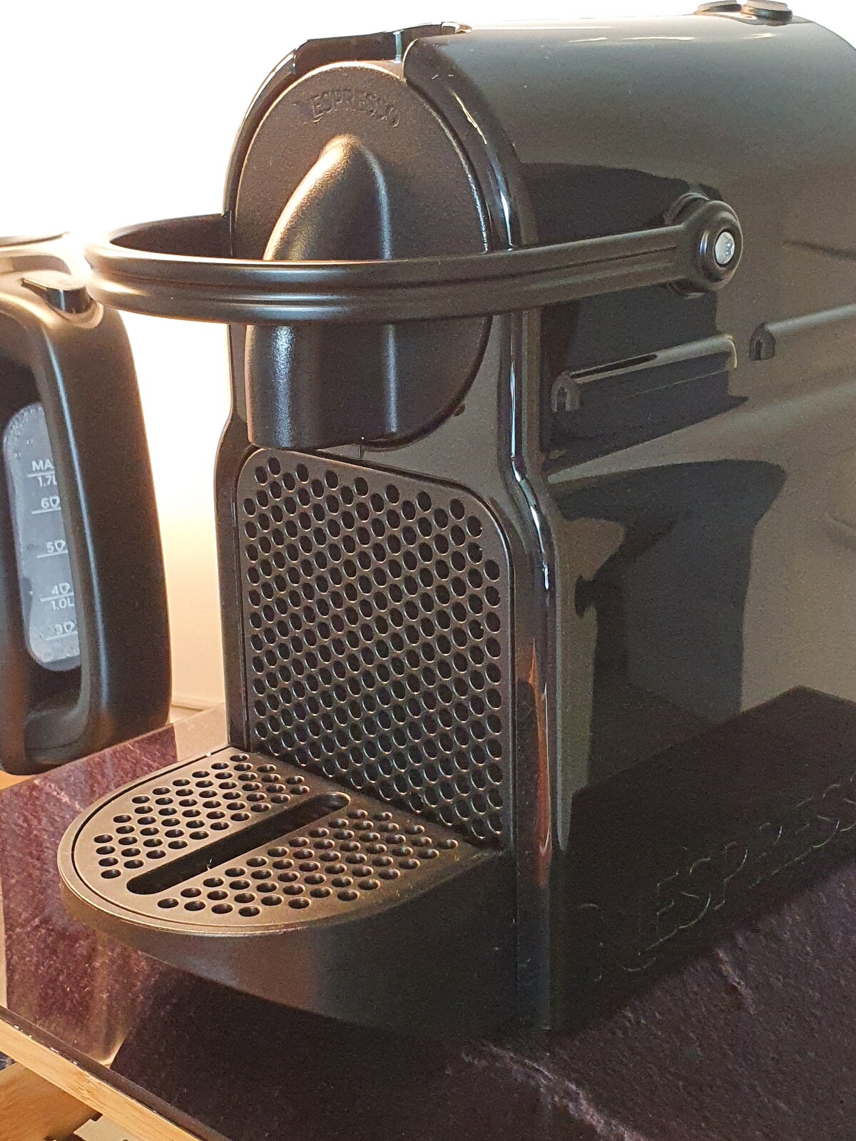 A sleek Nespresso coffee machine is positioned on a countertop, showcasing a polished black exterior. The machine features a prominent spout and a textured front panel with a grid pattern. A transparent water reservoir alongside indicates the coffee-making capabilities available.
