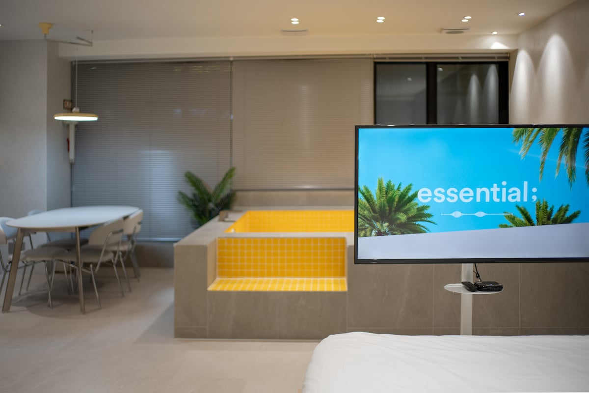 A modern living space is presented, featuring a wall-mounted television displaying the word "essential". A bright indoor pool with yellow tiles is visible nearby, alongside a round dining table with chairs and large windows covered with blinds.