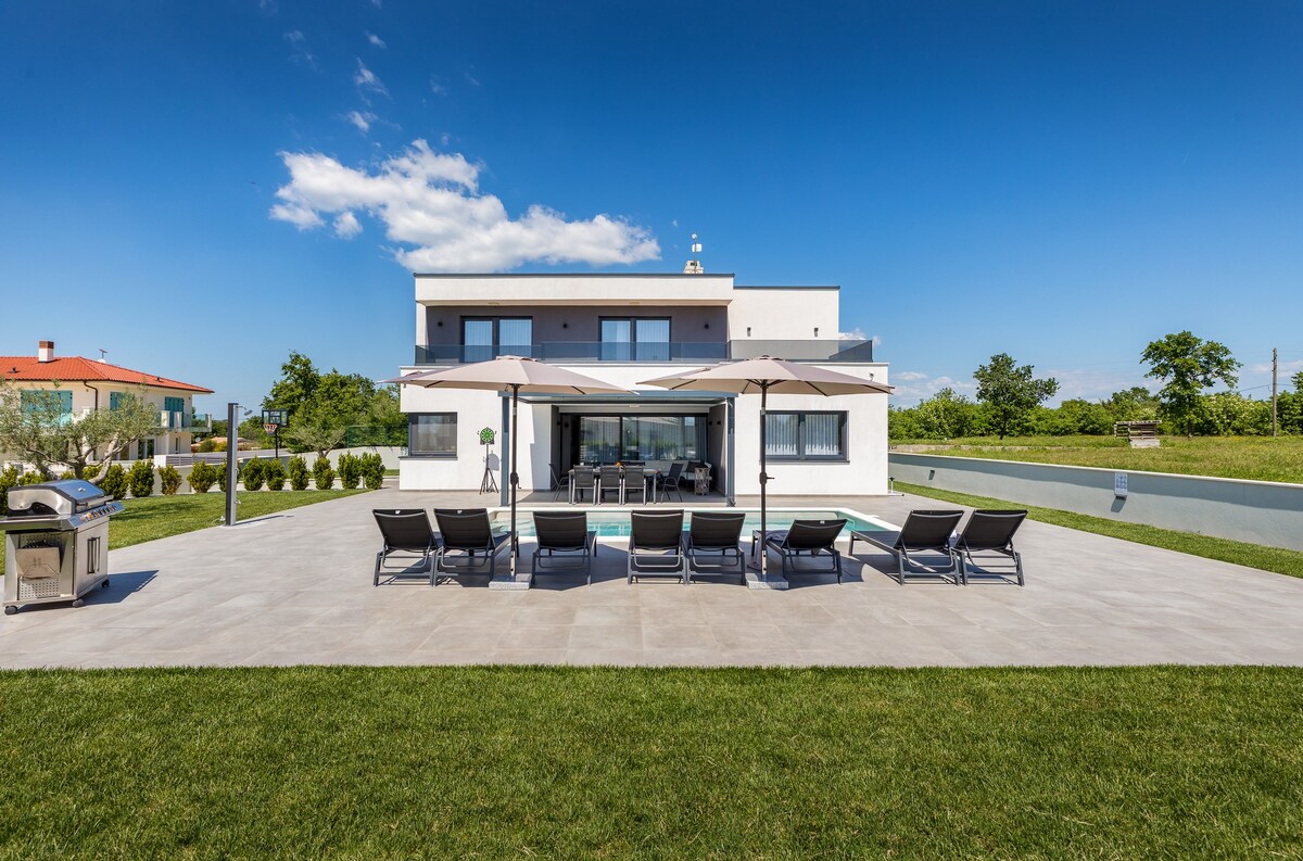 The villa's exterior is displayed, featuring a spacious terrace with eight sun loungers sheltered by elegant parasols. A saltwater pool sits at the center, framed by lush greenery. The modern two-story structure showcases large glass doors that connect the indoor living area to the outdoor space.