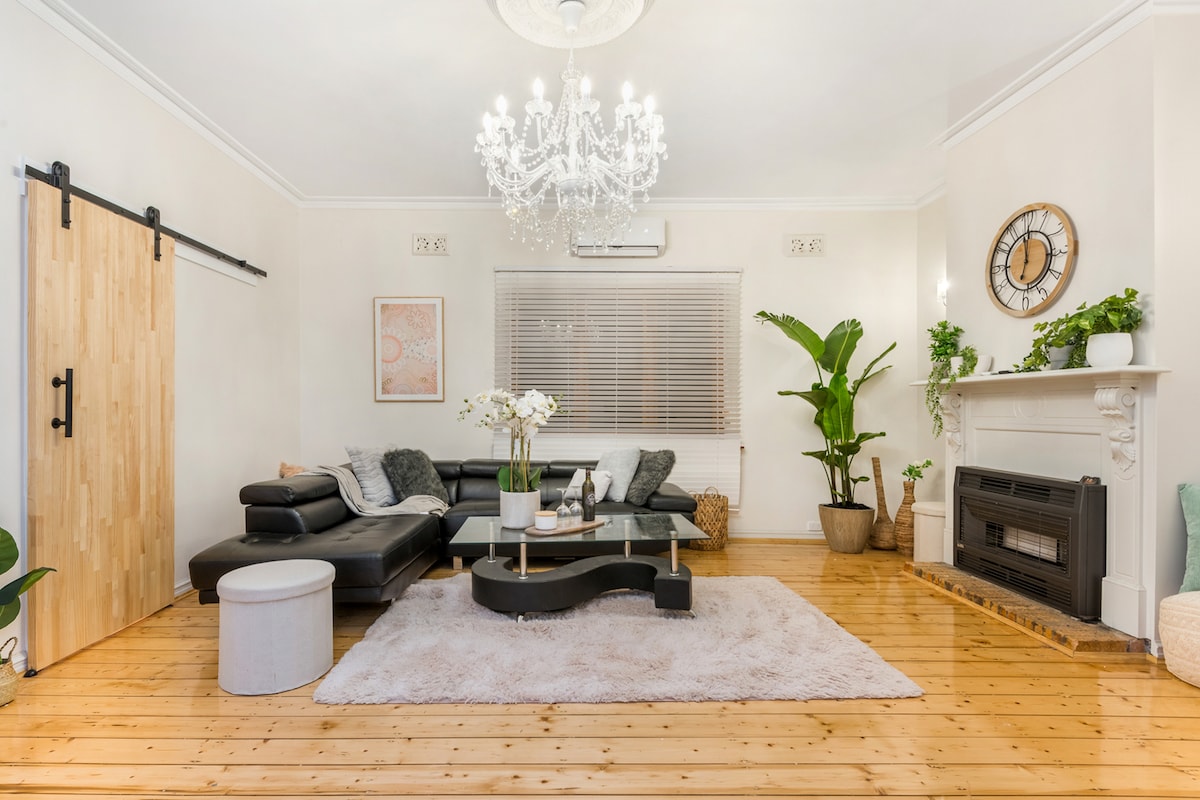 A light-filled living area features a stylish black sofa and a chic glass coffee table. A crystal chandelier hangs above, while decorative plants add a touch of greenery. Warm timber flooring complements the elegant ambiance of the room, showcasing a cozy and inviting environment.