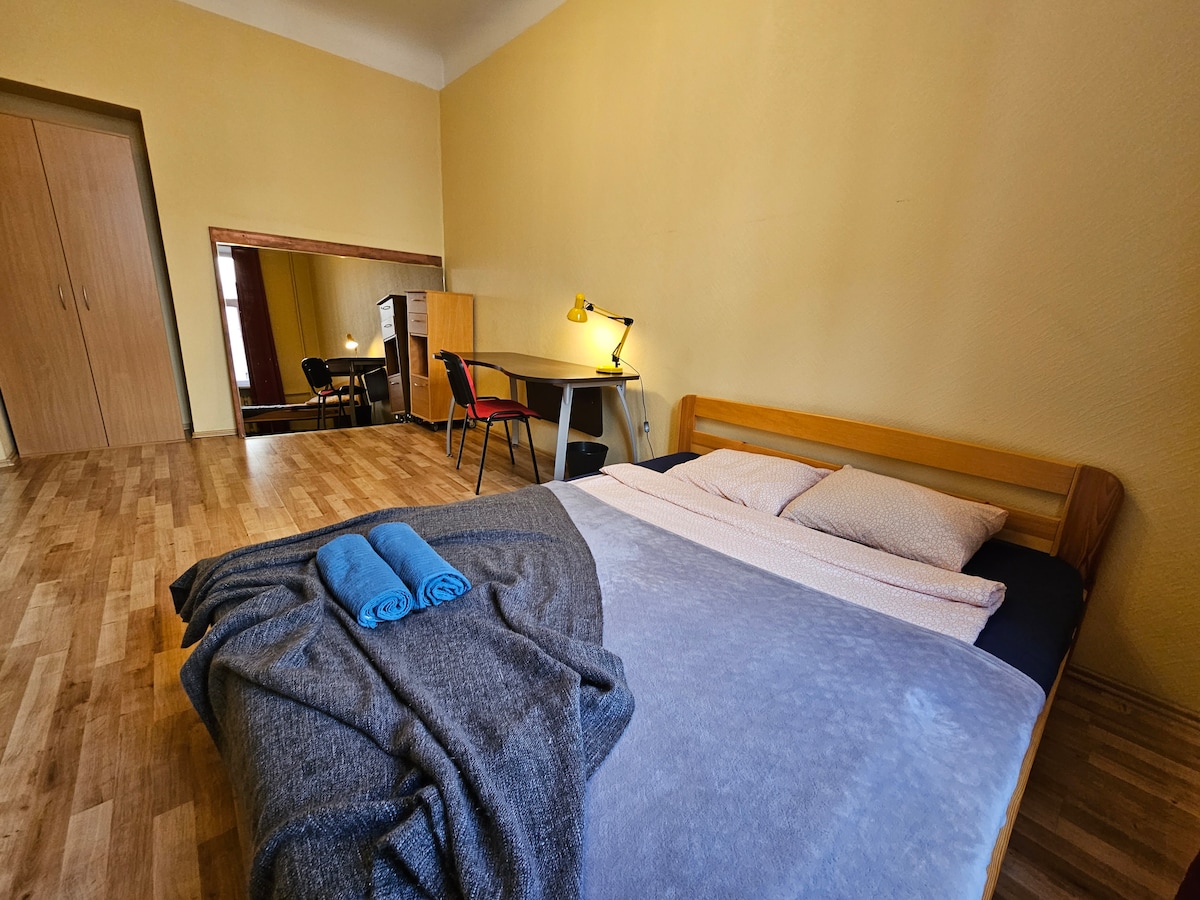 A spacious sleeping area features a large bed dressed in soft linens, flanked by plush pillows. A cozy throw blanket rests at the foot of the bed. A mirror reflects the room's warm wall color and wooden flooring, while a table and chairs are visible in the background.