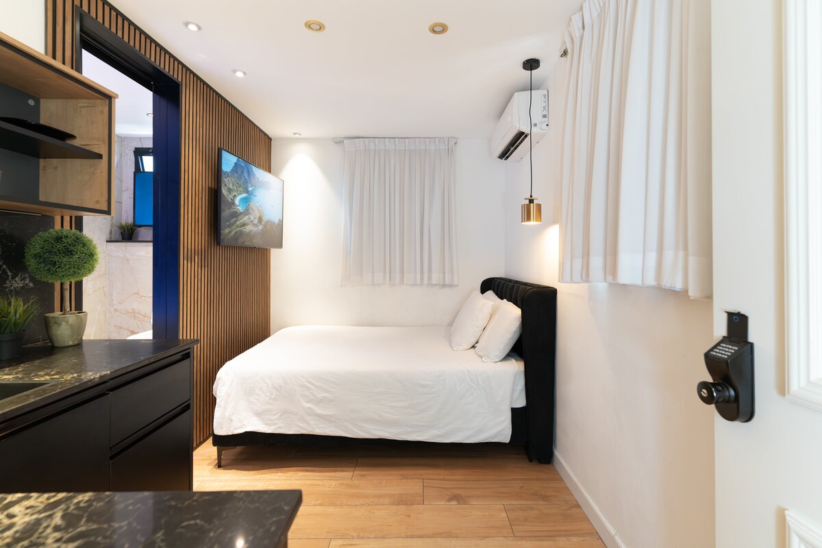 A well-lit studio apartment features a minimalist design with light tones. A spacious bed is positioned against a wall with wooden paneling, complemented by soft white bedding. A flat-screen TV is mounted on the wall, and sheer white curtains allow natural light to filter in.