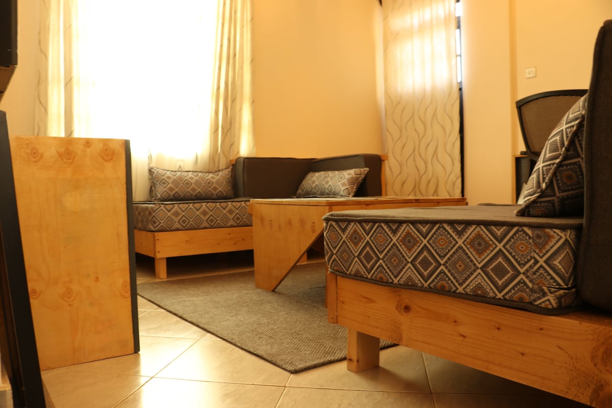 A comfortable living area features two wooden-framed sofas adorned with patterned cushions, positioned around a wooden coffee table. Natural light enters through sheer curtains, enhancing the warm tones of the space, while a textured area rug adds a cozy touch underfoot.