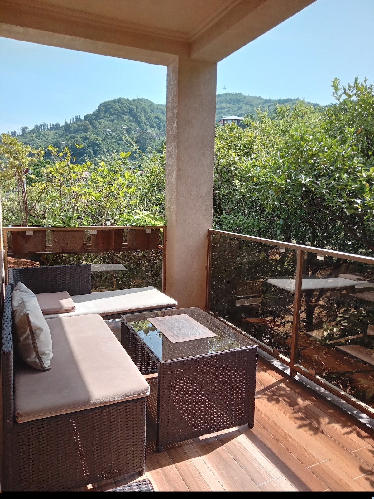 A shaded balcony is furnished with comfortable wicker seating and a glass-topped table. Lush greenery surrounds the balcony, offering a view of the nearby mountains and enhancing the tranquil outdoor experience.