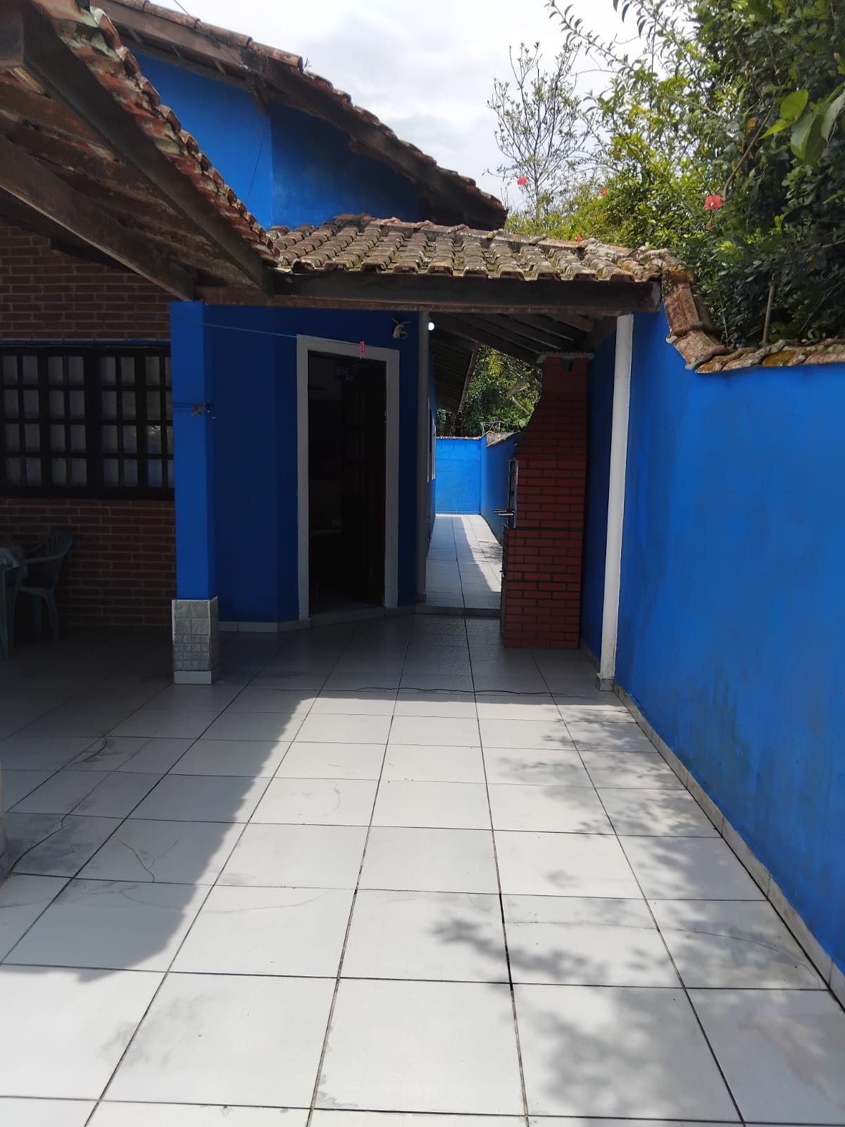 A bright blue pathway leads through the property, flanked by greenery and walls of blue paint. The tiled floor reflects natural light, creating an open and spacious atmosphere. An entrance can be seen at the end, guiding guests toward the interior of the house.