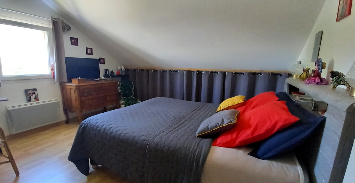 A cozy bedroom features a spacious bed adorned with bright pillows in red, yellow, and grey hues. Light enters through a nearby window, illuminating the wooden accents and decor. A dresser stands against the wall, enhancing the room's welcoming ambiance.