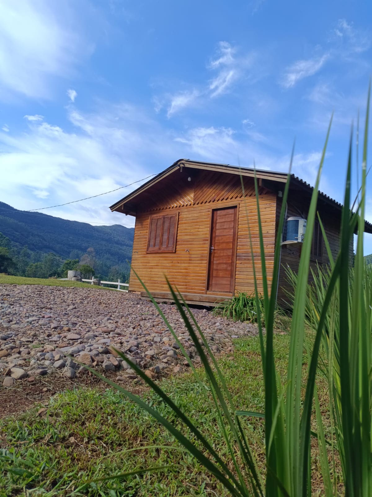 A wooden chalet is positioned against a backdrop of rolling hills and a clear blue sky. The structure features a brown door and wooden shutters. Lush green grass and pebbled ground surround the chalet, contributing to the natural setting.