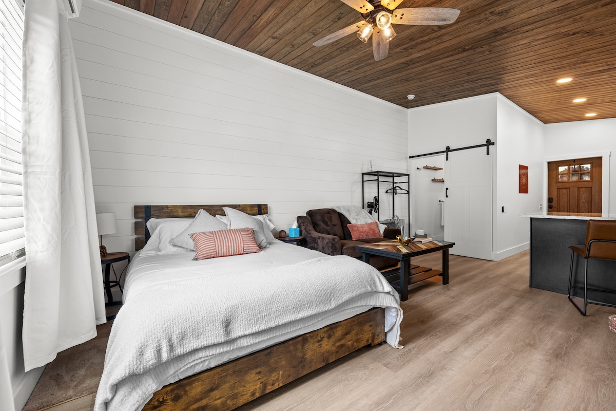 A spacious sleeping area features a wooden bed frame with a white comforter. A cozy armchair and a small coffee table sit nearby, while a clothing rack offers additional storage. Natural light floods the room through a large window with sheer curtains and a wooden ceiling adds warmth.