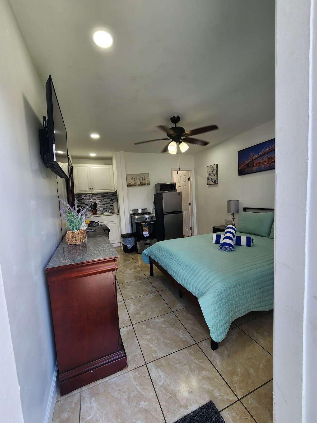 A compact room is visible, featuring a spacious bed with a light blue coverlet. A small kitchen area is included, with a refrigerator and microwave. Light fixtures provide bright illumination, and a ceiling fan is installed. A flat-screen TV is mounted on the wall, enhancing the functionality.