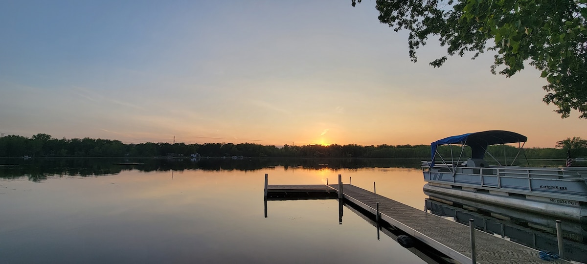 A tranquil lakeside scene is captured during sunset, with warm hues reflecting on the calm water surface. A wooden dock extends into the lake, alongside a pontoon boat anchored nearby, framed by lush greenery in the background.