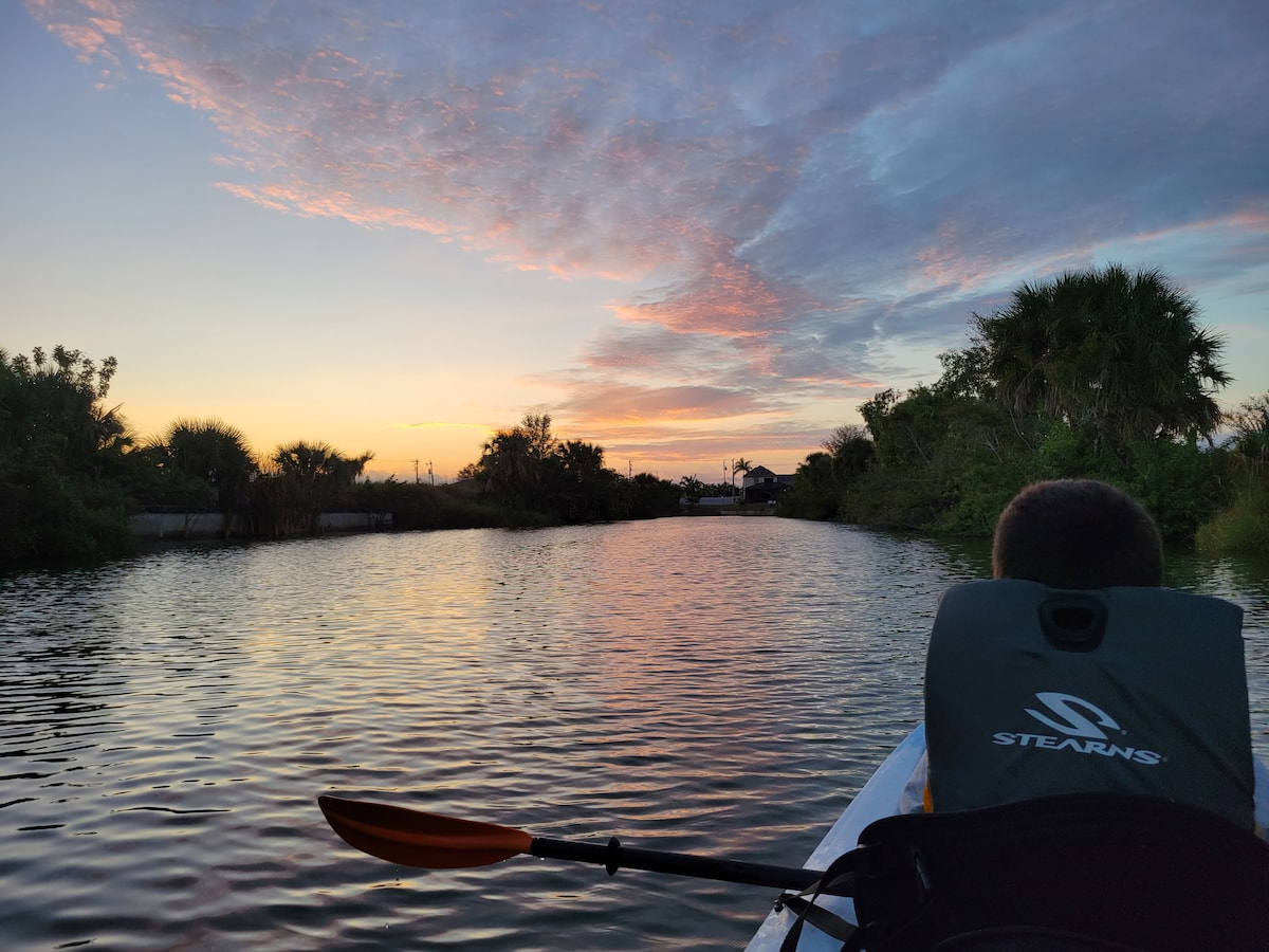 A tranquil scene features calm waters reflecting a colorful sunset sky with pink and orange hues. Lush greenery lines the waterfront, and a kayak is captured from the rear, showcasing a paddler wearing a life vest against the serene backdrop.
