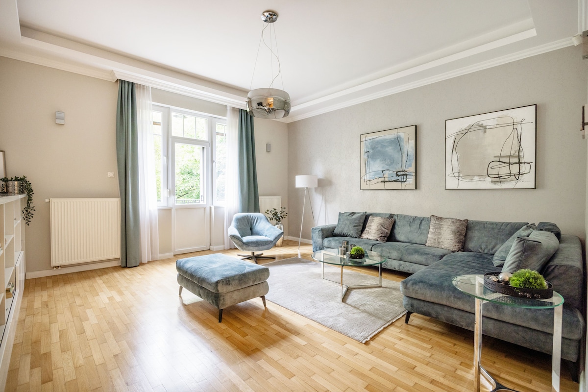 The living room features a large, comfortable sofa arranged around a coffee table. A stylish armchair and an ottoman accompany the seating area. Natural light fills the space through large windows with sheer curtains, illuminating contemporary artwork on the walls.