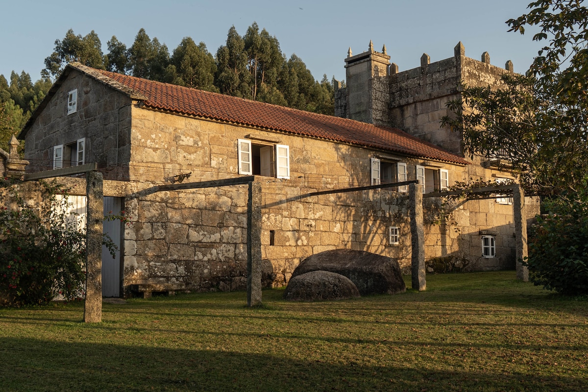 The exterior of a rustic stone house is framed by lush greenery and a well-kept lawn. White windows contrast with the earthy tones of the walls. An old stone wall and unique rock feature add character to the setting, suggesting a serene and historic ambiance.