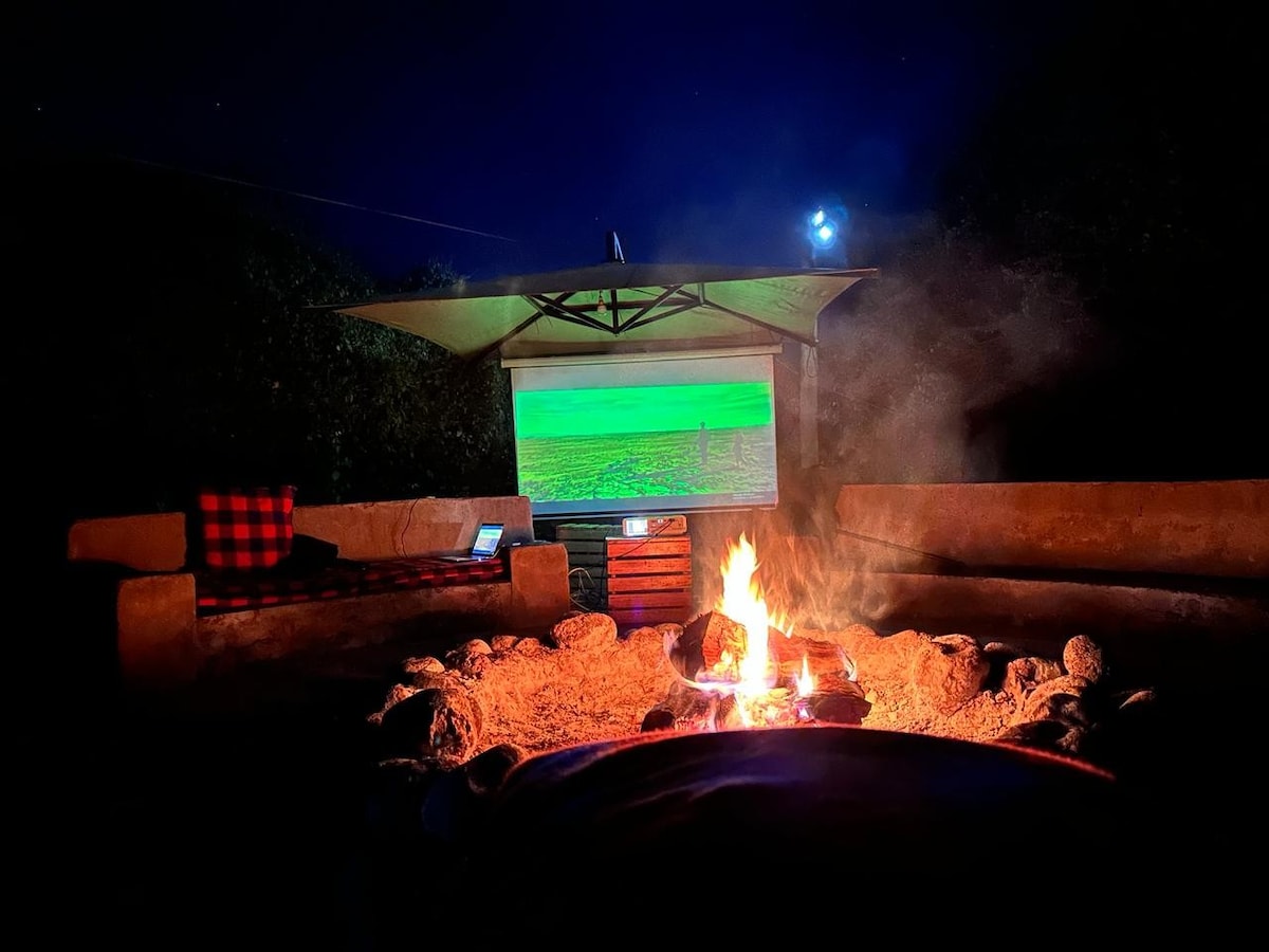 An outdoor seating area is arranged around a circular fire pit, with comfortable benches visible nearby. A screen is mounted overhead, displaying a scenic view. The night sky is illuminated by a bright moon, creating a serene atmosphere.