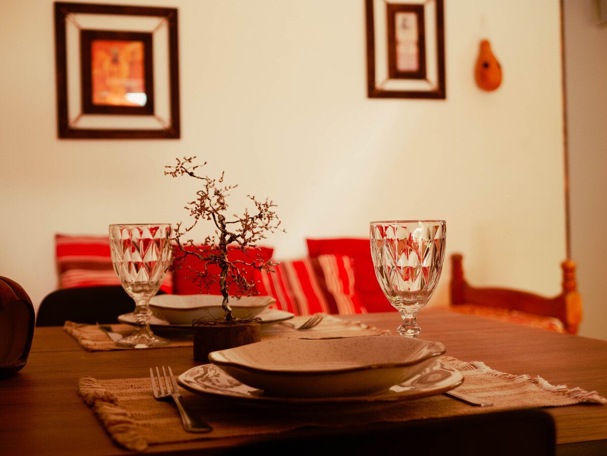A dining area is presented with a wooden table set for a meal. Glassware and plates are arranged elegantly with a decorative centerpiece. The walls are adorned with framed art, and a cozy seating arrangement is visible in the background.