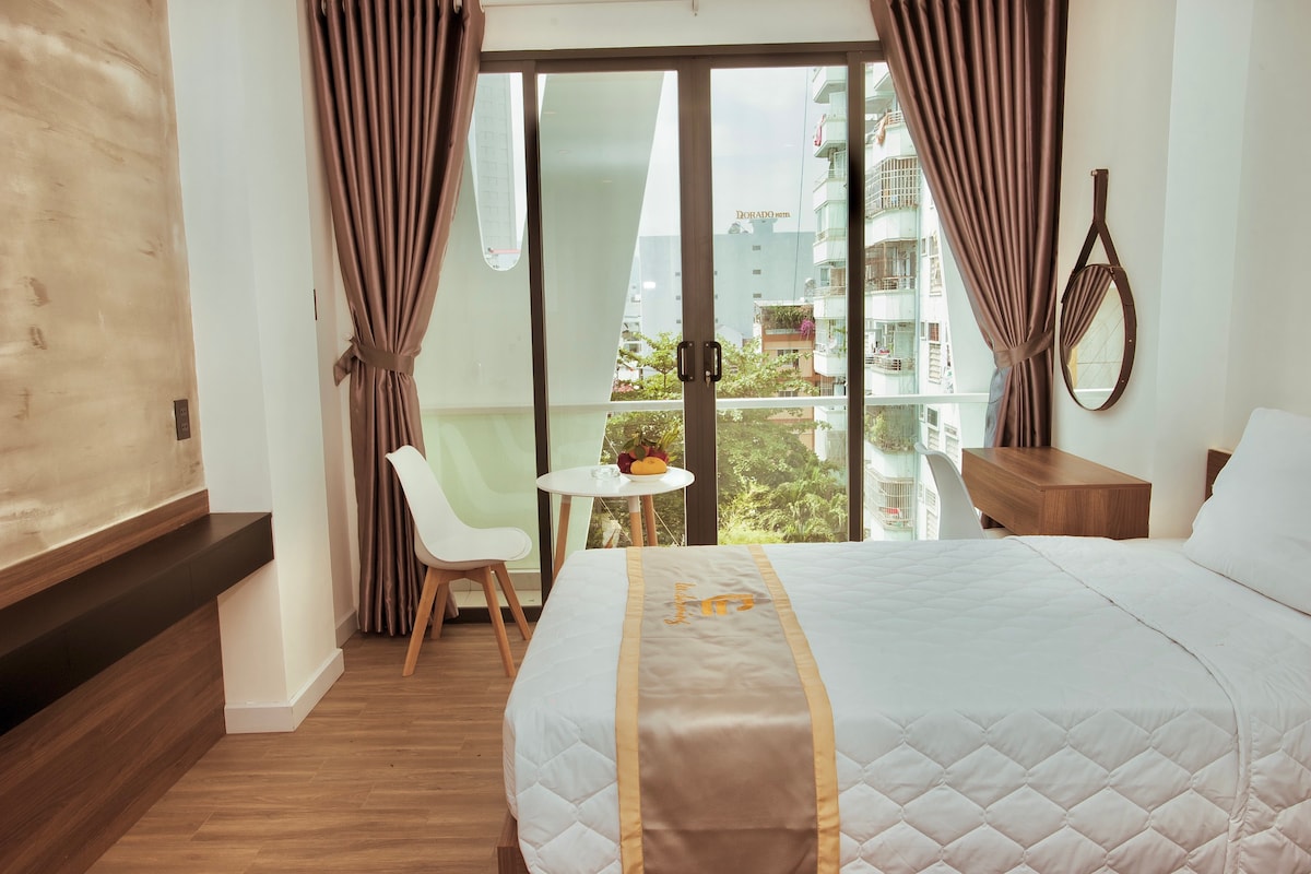 A bright room features a spacious bed dressed in neutral linens. Large glass doors open to a balcony, allowing natural light to fill the space. A round table with two chairs is positioned beside the window, offering a view of the surrounding area.