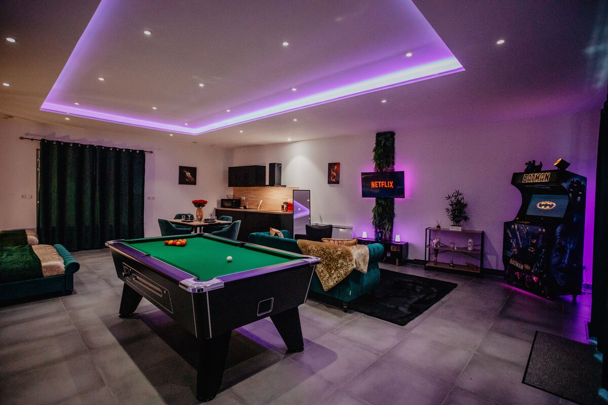An open living area features a green billiard table as a central focus, complemented by a sleek black arcade machine themed around Batman. Soft lighting accents the space, while a large TV is mounted on the wall, displaying the Netflix logo.