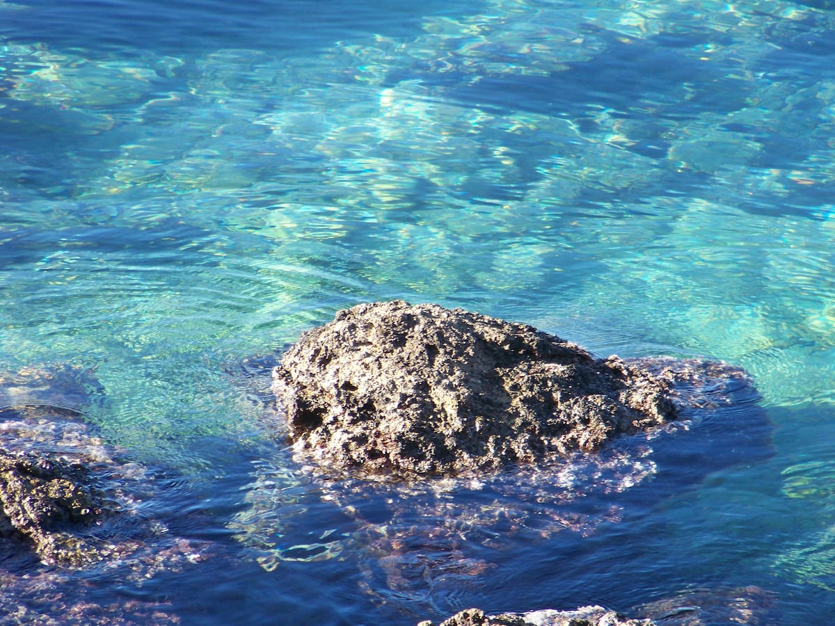 Clear turquoise waters gently lap against a rocky shoreline, showcasing a large stone partially submerged. Sunlight reflects off the surface, creating patterns of light that dance in the water. The scene conveys a serene coastal environment, typical of the area near the beach.