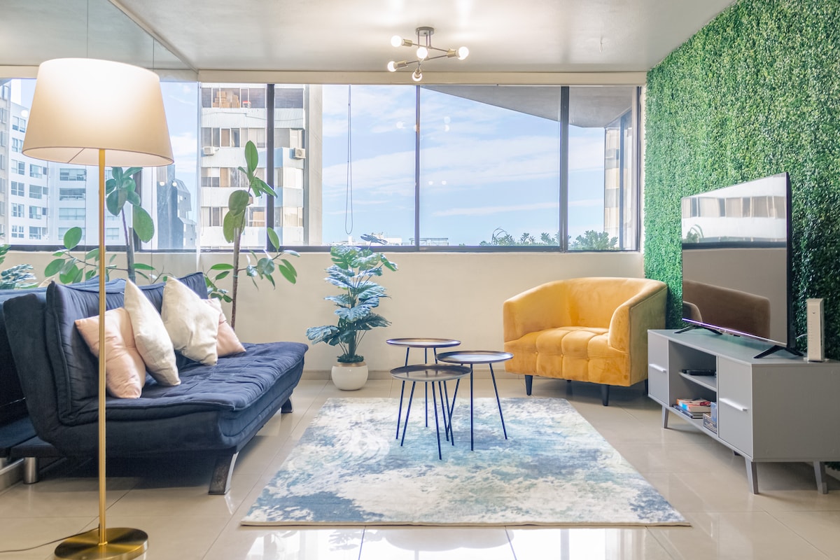 A cozy living area features a blue sofa bed, a yellow armchair, and a coffee table set against large windows offering city views. A rug with abstract designs and a potted plant enhance the space, complemented by a standing lamp and a wall adorned with greenery.