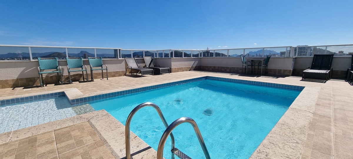 A rooftop area features a refreshing swimming pool surrounded by a textured stone deck. Comfortable lounge chairs and a small dining table with chairs are positioned nearby, offering views of the horizon and cityscape under a clear blue sky.