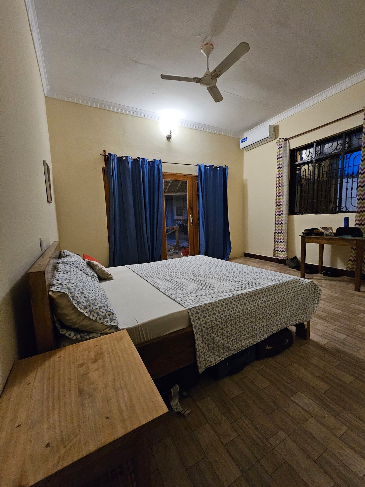 A spacious room features a comfortable bed covered with a patterned blanket. Blue curtains frame a window that opens to a view outside. A wooden table is positioned nearby, and a fan is mounted on the ceiling. Wood-toned flooring complements the warm decor.