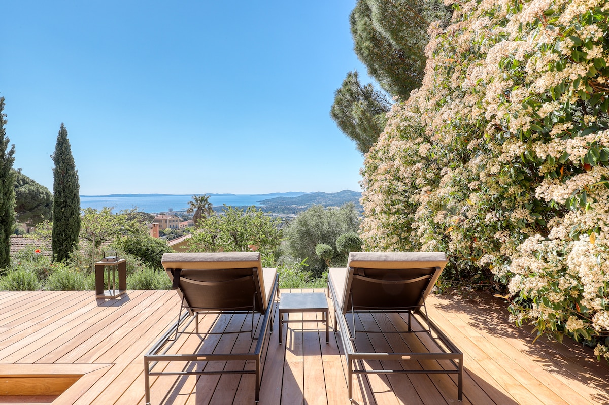 A sun-soaked terrace features two lounge chairs facing a scenic view of the sea. Surrounding vibrant greenery creates a serene environment, while distant vistas of hills and houses can be seen under a clear blue sky.