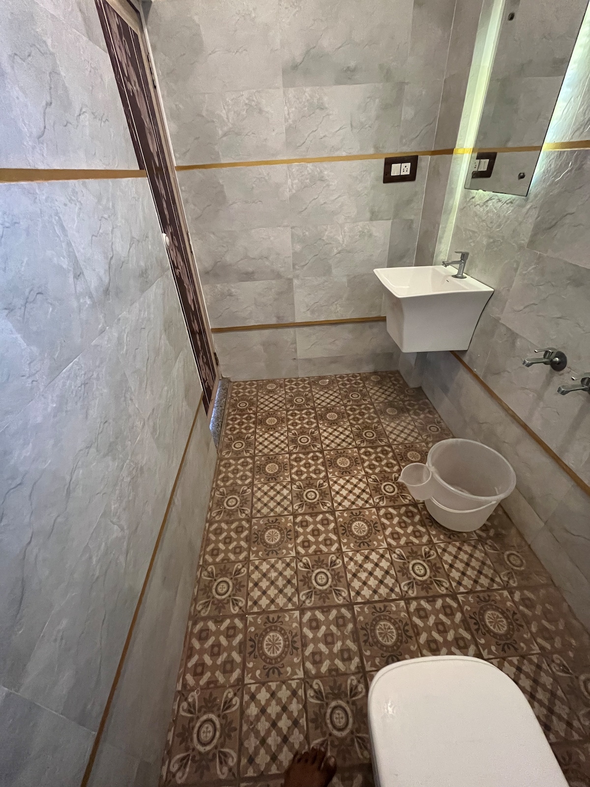 The modern bathroom features tiled walls and a decorative floor. A white sink is mounted against the wall, with plumbing fixtures nearby. A bucket sits on the floor, and light enters from a narrow window, creating a well-lit space.