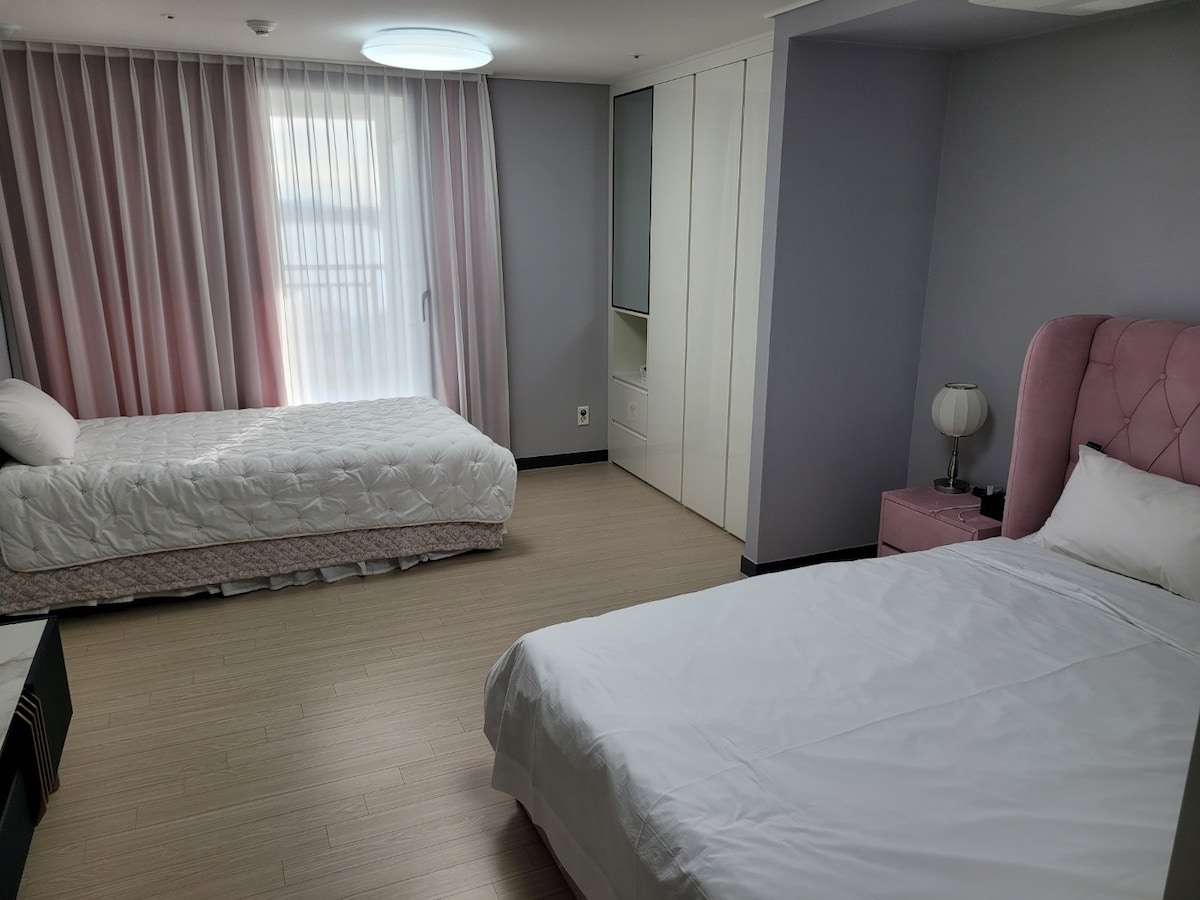 A bright, minimalist guest room features two spacious beds, one with a soft pink headboard. Light streams through large curtains, and modern furniture complements the calming color scheme of the walls. A bedside lamp and wardrobe enhance the room's functionality.
