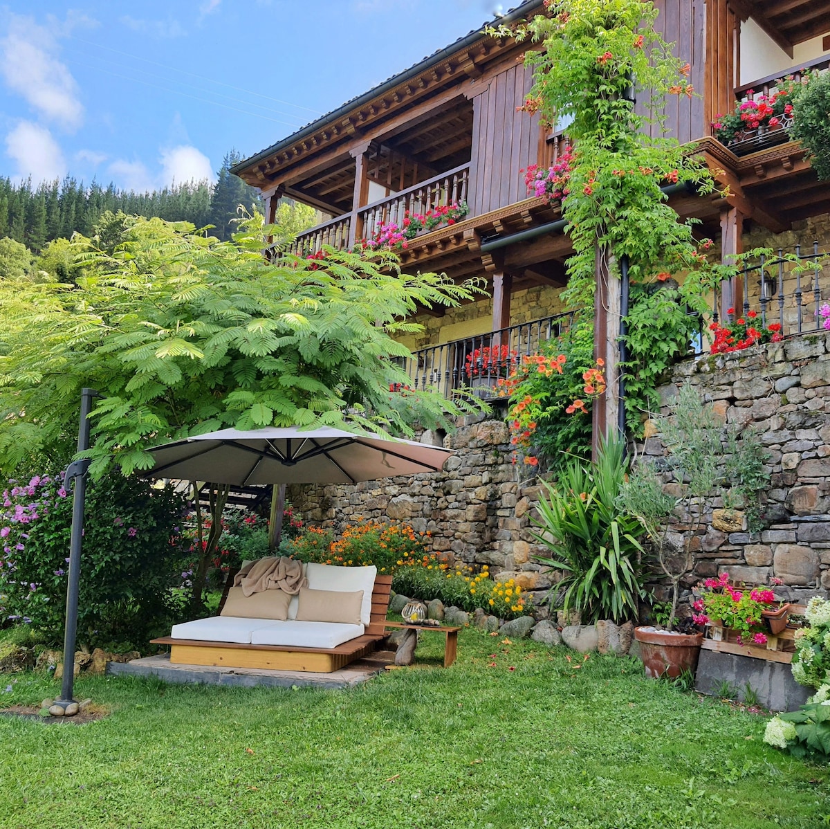 A charming outdoor seating area is positioned under a large canopy, featuring a spacious, cushioned lounge chair. Lush greenery and vibrant flowers surround the space, while a rustic stone wall provides a natural backdrop to the inviting setting.