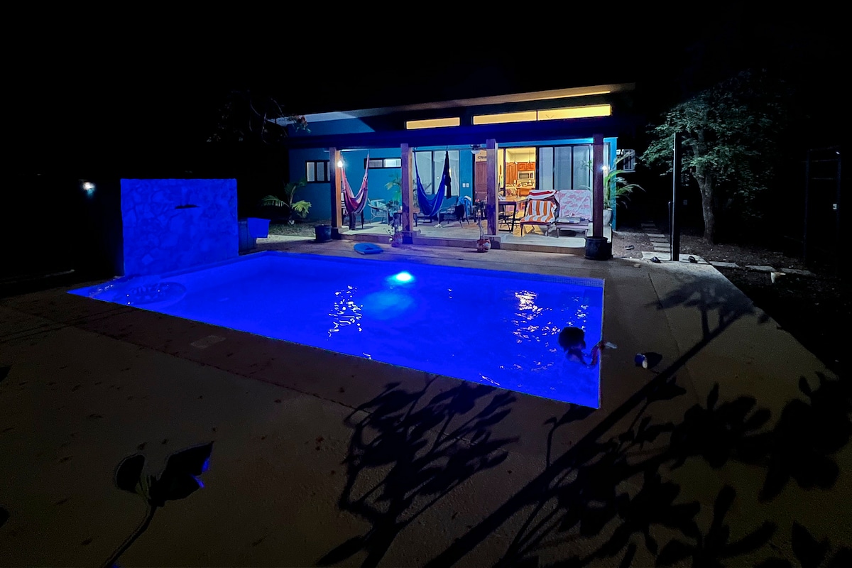 A serene nighttime view of a private pool is highlighted by soft blue lighting, reflecting on the water's surface. Hammocks are draped nearby, and the illuminated interior of the house can be seen through expansive glass doors, creating a peaceful ambiance.