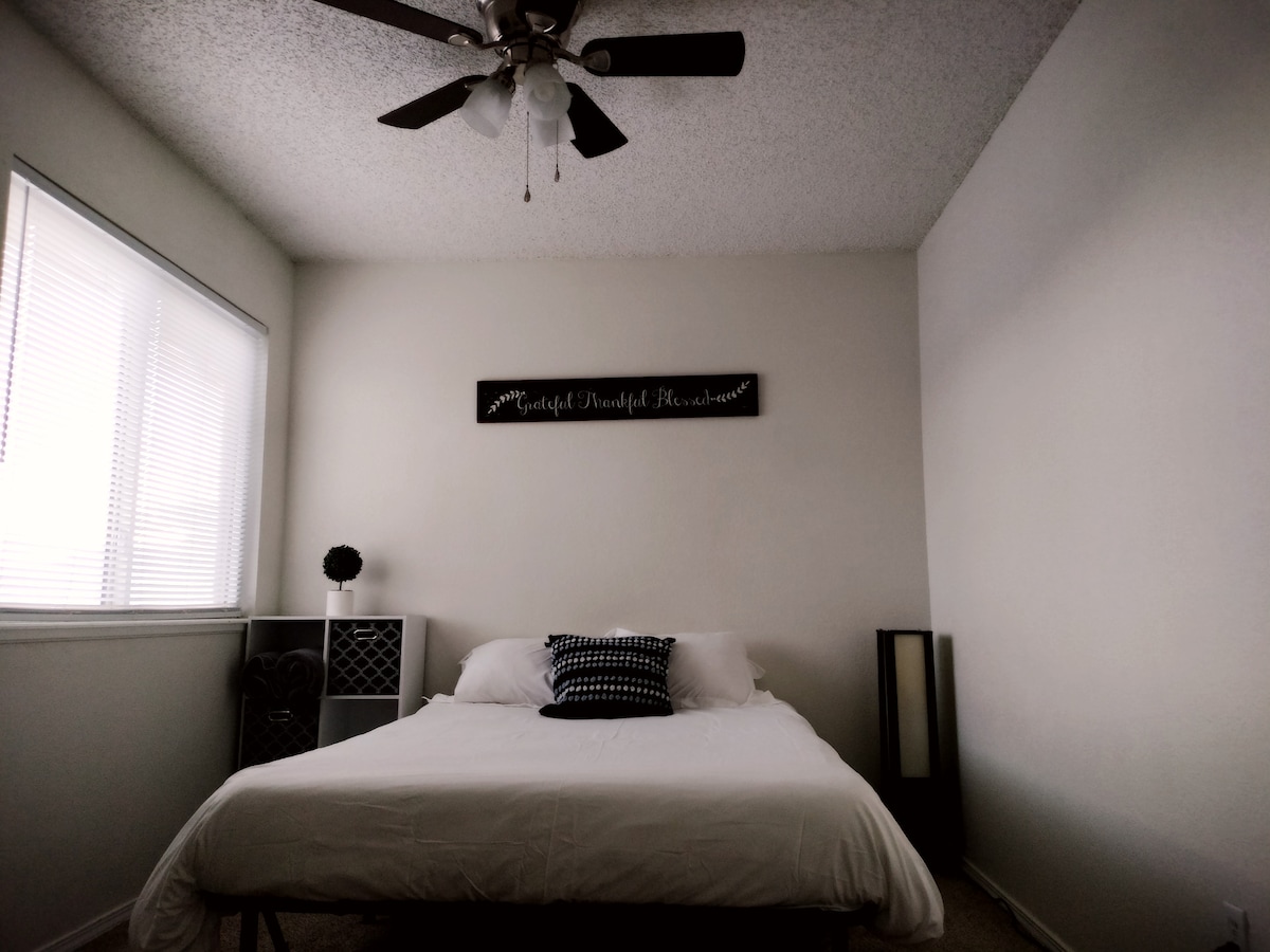 A bright bedroom features a spacious bed with a white bedding set. A decorative wall sign is mounted above the bed, with natural light filtering through the window. A nightstand and a full-length mirror are positioned in the room, contributing to a clean and simple aesthetic.