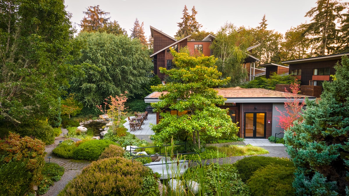 The landscaped garden features a variety of lush greenery and colorful plants, creating a serene environment. A pathway leads to a peaceful patio area furnished with seating, surrounded by water features and vibrant flora, providing a tranquil backdrop for relaxation.