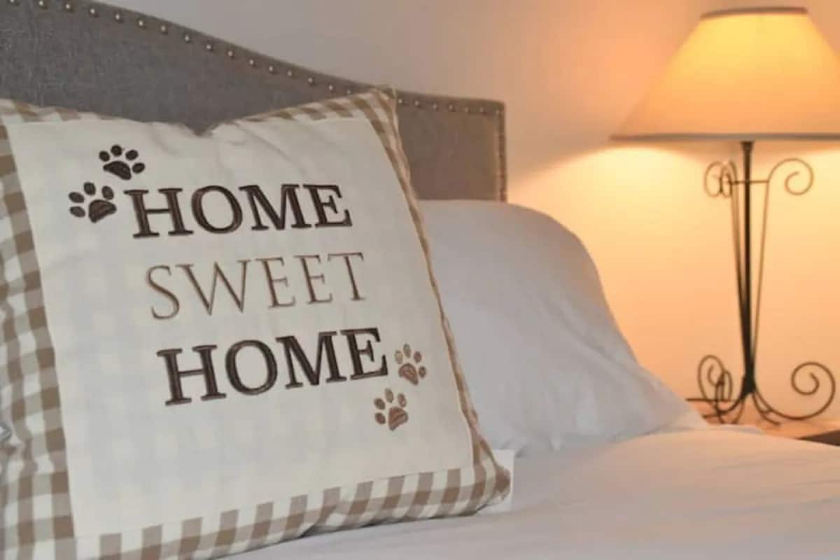 A decorative pillow with the phrase 'Home Sweet Home' is placed on a spacious bed. A bedside lamp casts a warm light, creating a cozy ambiance, while a soft white blanket is neatly arranged on the bed.